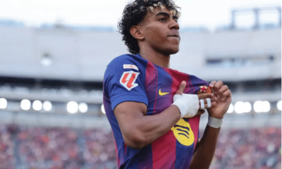 A mid-shot of Lamine Yamal on the pitch, with the stands blurred behind him. His right hand is bandaged with buddy strapping on his middle and fourth fingers. That hand crosses his torso to touch his chest