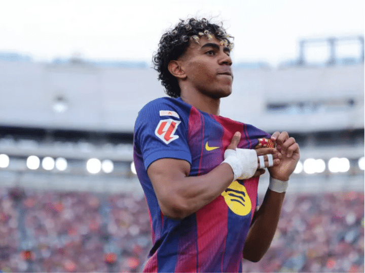 A mid-shot of Lamine Yamal on the pitch, with the stands blurred behind him. His right hand is bandaged with buddy strapping on his middle and fourth fingers. That hand crosses his torso to touch his chest