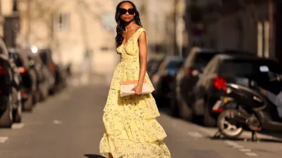 Emilie Joseph @in_fashionwetrust wears black sunglasses, a pale yellow with green and yellow flower print pattern V-neck / tank-top / backless / belted / ruffled maxi long dress, a beige fabric ruffled clutch, silver rings, pale yellow matte leather pointed pumps heels shoes, during a street style fashion photo session, on March 26, 2022 in Paris, France.