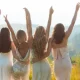 Rear view of female friends in elegant dresses looking at the mountains, embracing and raising arms at sunset. Young women during bachelorette party.