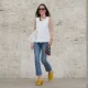 Alba Garavito Torre wears black sunglasses, long straight brown hair with a center part, a white pleated peplum sleeveless blouse by Commense, and blue high-rise raw-hem denim jeans. A yellow clog sandal with floral detailing by AGL is worn, paired with a jean denim and brown mini Jackie handbag by Gucci. Accessories include a chunky necklace with colorful natural stones by Sempiterno Paris and gold jewelry. Full body shot, during a street style fashion photo session, on May 02, 2025 in Paris, France.