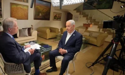 Israeli Prime Minister Benjamin Netanyahu sits across from CBS News reporter Major Garrett in a living room setting with the standing lights shining on them