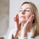 A focused adult woman practices facial self-massage in her well-lit bathroom. Using her fingertips, she gently smoothes her skin to improve circulation, reduce tension, and promote a healthy, natural glow. This authentic at-home wellness moment captures the popular beauty and self-care trend