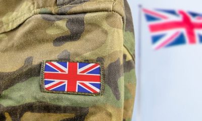 Close up of a soldier's arm in camo with a union flag badge. A union flag is flying in the background. Defence Universities Alliance