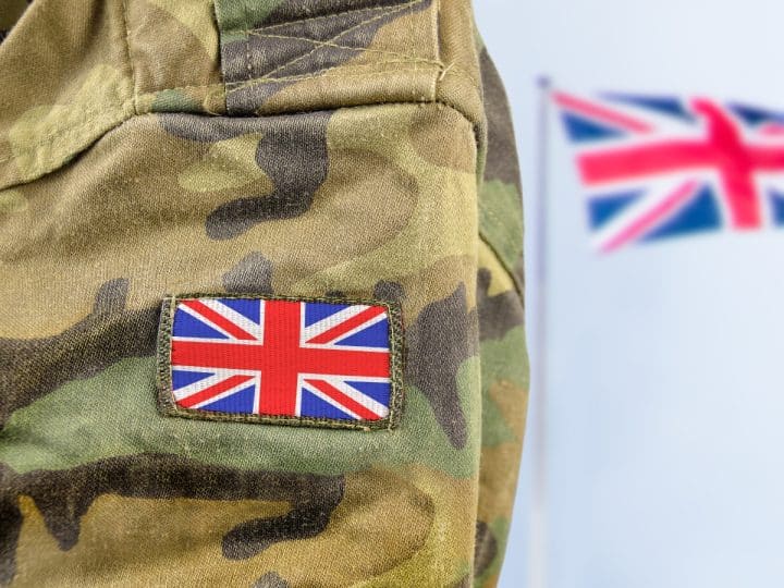 Close up of a soldier's arm in camo with a union flag badge. A union flag is flying in the background. Defence Universities Alliance
