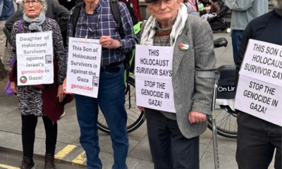 Holocaust survivor Stephen Kapos (right) and descendants of Holocaust survivors take part in an April 13, 2024 London protest against the Gaza genocide. He wears a flat cap and wool jumper underneath a wool blazer with one arm in a walking stick with arm support. On his torso is a poster that reads: ‘This Holocaust survivor says stop the genocide in Gaza’.