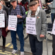 Holocaust survivor Stephen Kapos (right) and descendants of Holocaust survivors take part in an April 13, 2024 London protest against the Gaza genocide. He wears a flat cap and wool jumper underneath a wool blazer with one arm in a walking stick with arm support. On his torso is a poster that reads: ‘This Holocaust survivor says stop the genocide in Gaza’.