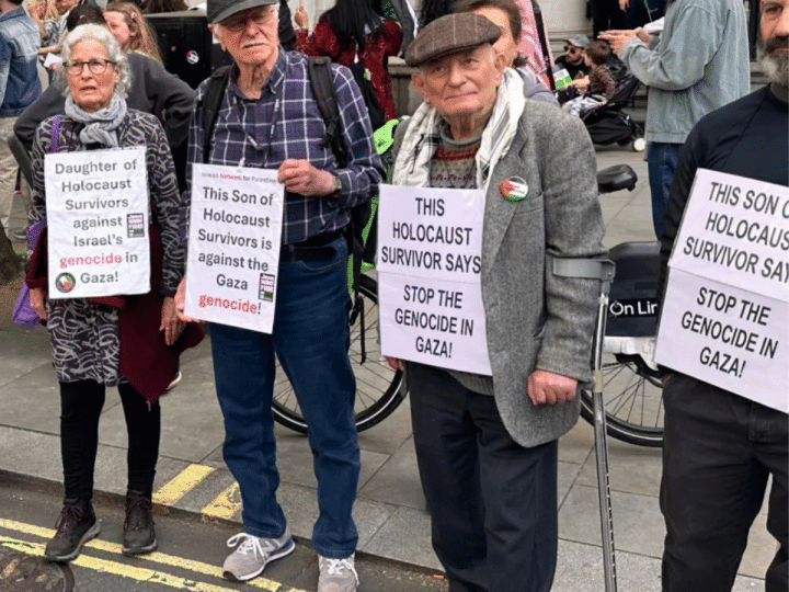 Holocaust survivor Stephen Kapos (right) and descendants of Holocaust survivors take part in an April 13, 2024 London protest against the Gaza genocide. He wears a flat cap and wool jumper underneath a wool blazer with one arm in a walking stick with arm support. On his torso is a poster that reads: ‘This Holocaust survivor says stop the genocide in Gaza’.