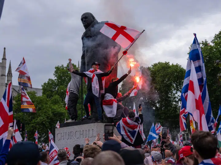 People, largely white men, sit on a Winston Churchill statue with flares and wearing Union Jack, England and Scotland flags as they take part in the rally where Muslims experienced hate speech