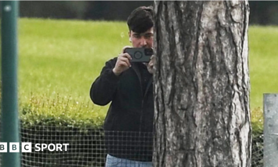 A man in a dark jacket and blue jeans stands partially concealed by a tree trunk and holds a phone up in two hands as if filming and has a white earphone in his right ear
