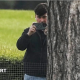 A man in a dark jacket and blue jeans stands partially concealed by a tree trunk and holds a phone up in two hands as if filming and has a white earphone in his right ear