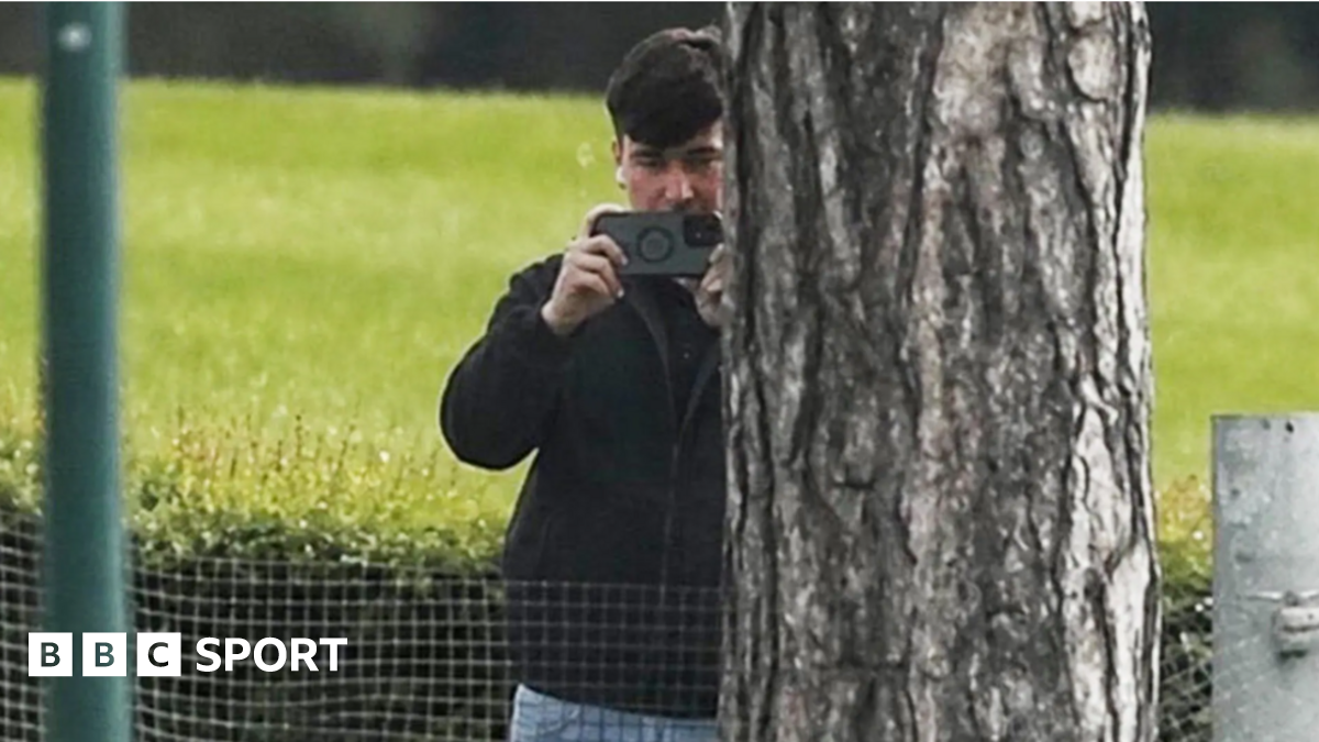 A man in a dark jacket and blue jeans stands partially concealed by a tree trunk and holds a phone up in two hands as if filming and has a white earphone in his right ear