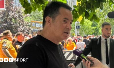 Hull City owner Acun Ilicali being interviewed outside Wembley Stadium, wearing a black t-shirt. Crowds on Olympic Way can be seen behind him.