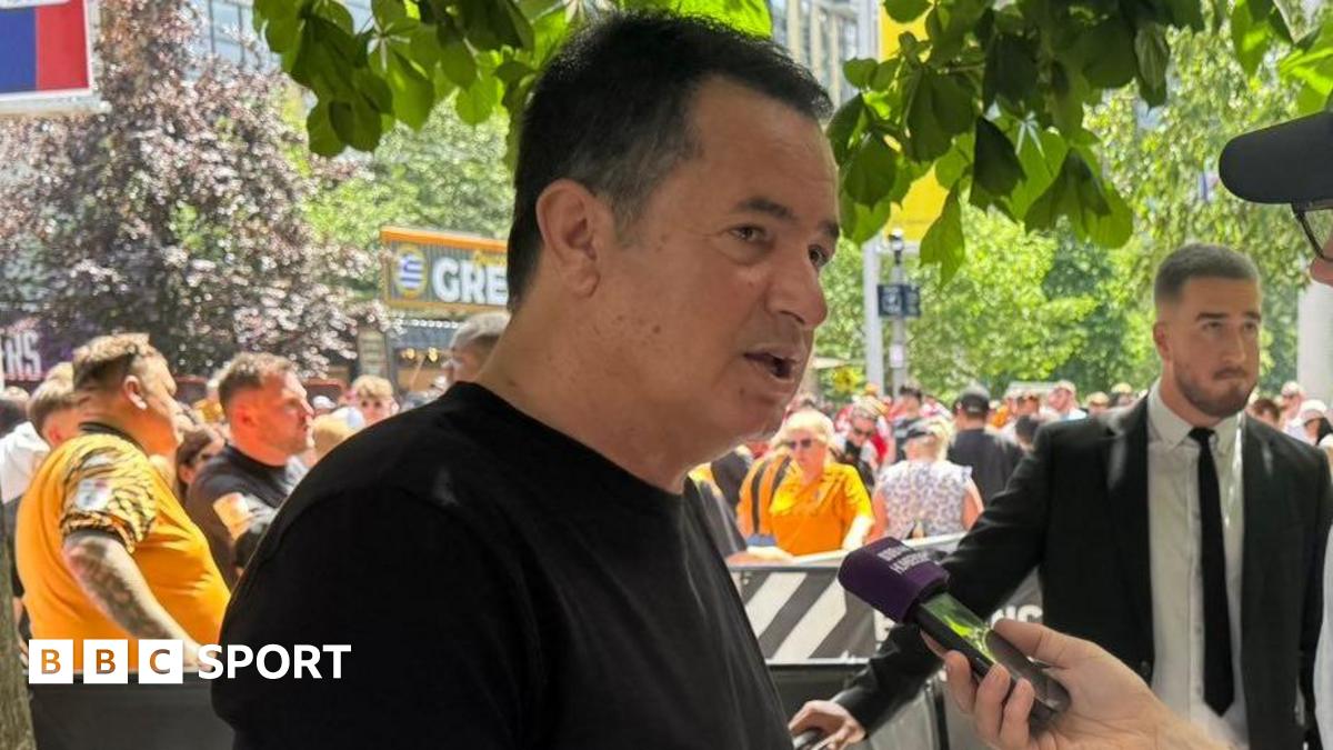 Hull City owner Acun Ilicali being interviewed outside Wembley Stadium, wearing a black t-shirt. Crowds on Olympic Way can be seen behind him.