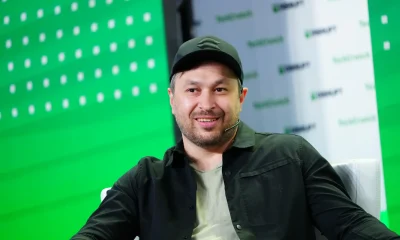 Smiling man in a black shirt and cap sitting on stage during a conference.