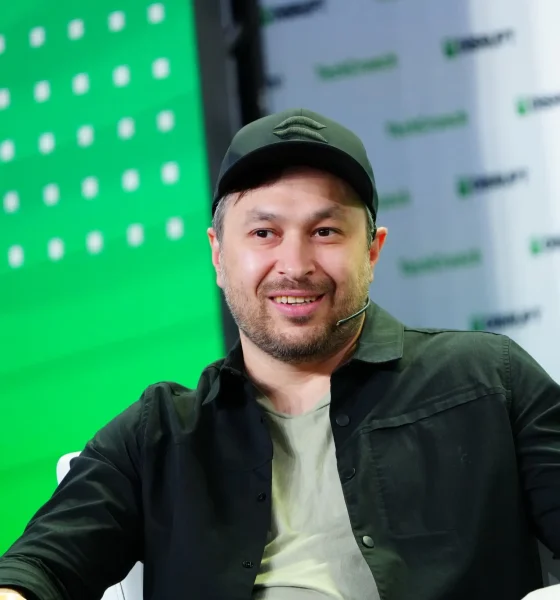 Smiling man in a black shirt and cap sitting on stage during a conference.
