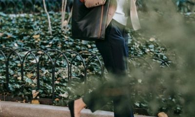Black woman walks outside on her way to work; she is wearing what could be some of the best work pants for women in 2024! She's carrying a tote bag and drinking a coffee while she walks behind a short circular fence
