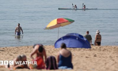 People on beach during May heatwave. One man wades in the sea whist two people paddle by in a kayak