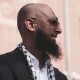 Majid Freeman, wearing sunglasses, smiles and looks into the distance. The stylish photograph captures his side profile.