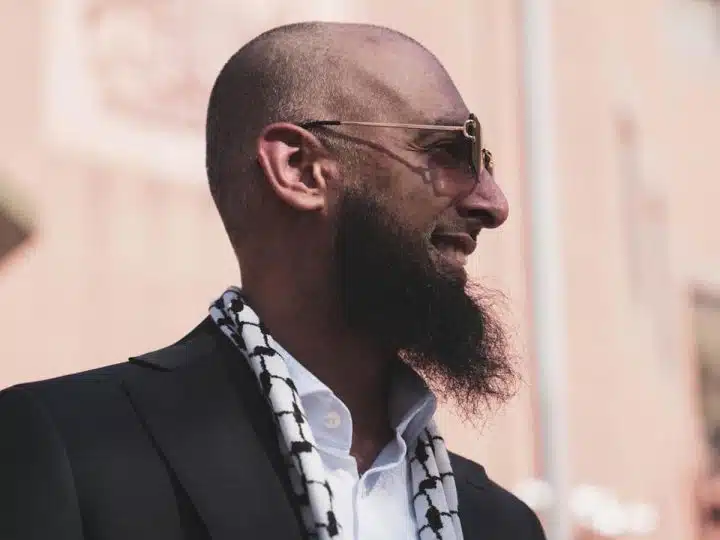 Majid Freeman, wearing sunglasses, smiles and looks into the distance. The stylish photograph captures his side profile.