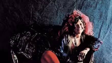 Janis Joplin with a pink headdress reclines on a couch and smiles on the album cover for Pearl (1971).