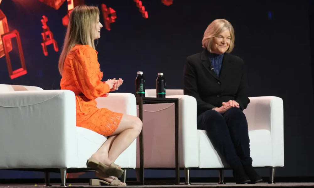 Two women sit on a stage during an event, one in an orange dress and the other in a black jacket.