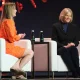 Two women sit on a stage during an event, one in an orange dress and the other in a black jacket.