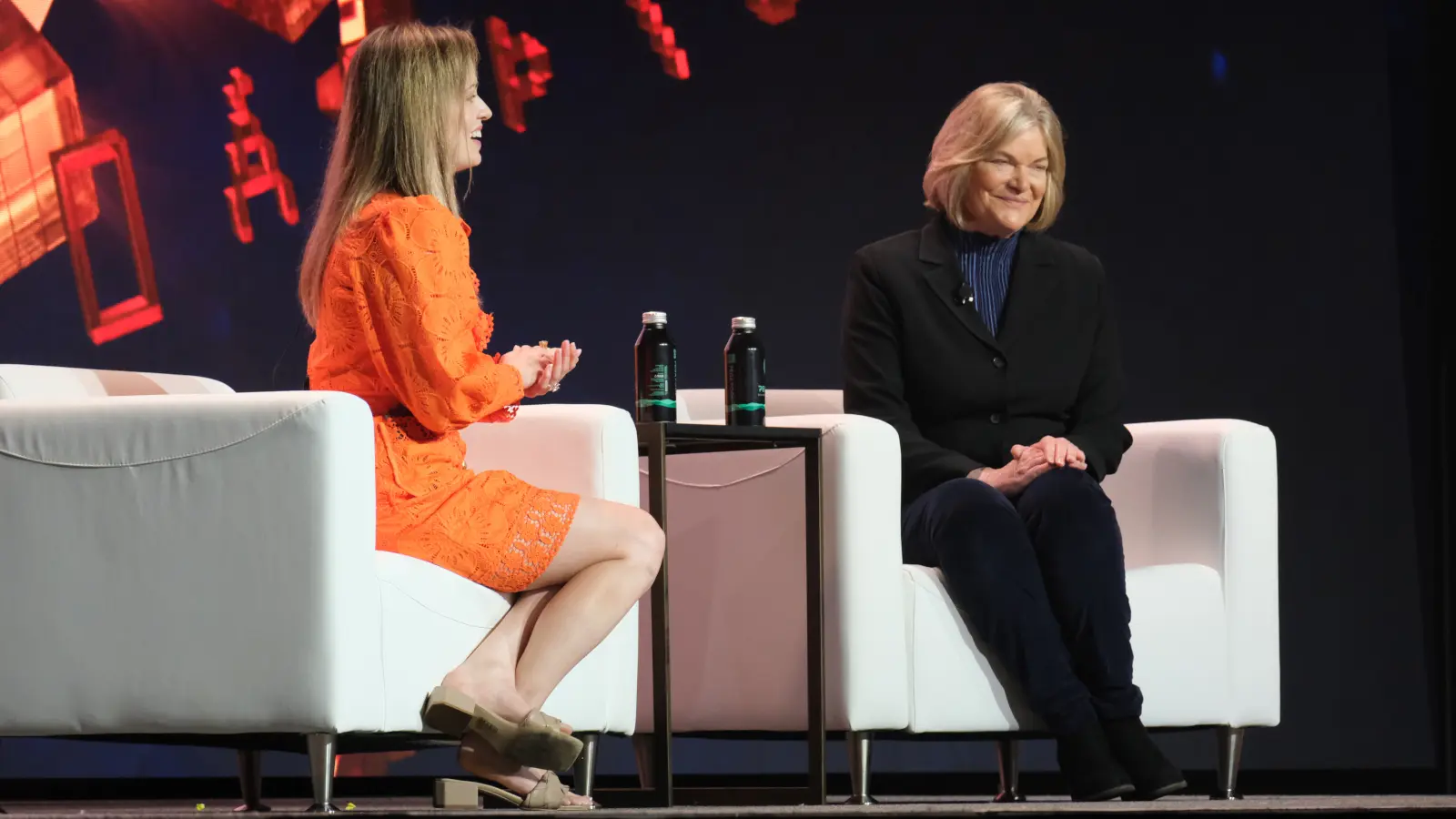 Two women sit on a stage during an event, one in an orange dress and the other in a black jacket.