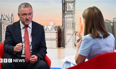 Housing, Communities and Local Government Secretary Steve Reed appearing on the Sunday with Laura Kuenssberg, with presenter Victoria Derbyshire standing in. The pair sit on red bucket chairs. Reed wears a black suit with a white shirt and a red tie. Derbyshire is wearing a pastel blue dress with short sleeves. She has shoulder length, dark blonde hair.
