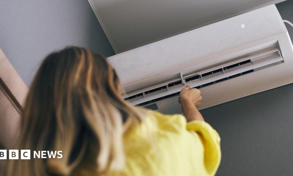 A woman with brown hair is wearing a yellow jumper and is pointing a control at a white air conditioned unit on a wall.