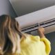 A woman with brown hair is wearing a yellow jumper and is pointing a control at a white air conditioned unit on a wall.