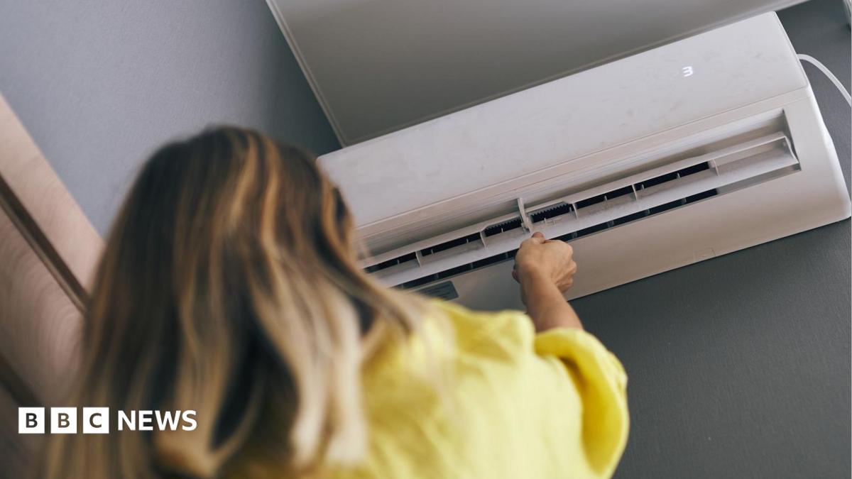 A woman with brown hair is wearing a yellow jumper and is pointing a control at a white air conditioned unit on a wall.