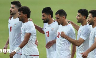 Six Iran players, wearing white Iran shirts, stand in a line