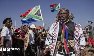 Hundreds of protesters marching outdoors, chanting and holding south african flags and signs