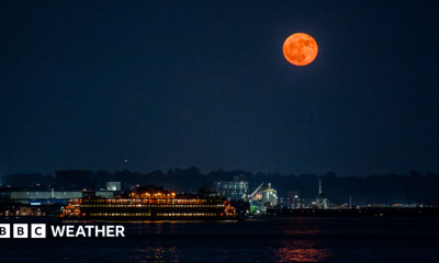 An orange-hued moon hangs in the sky over a river illuminated by lights from nearby properties