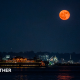 An orange-hued moon hangs in the sky over a river illuminated by lights from nearby properties