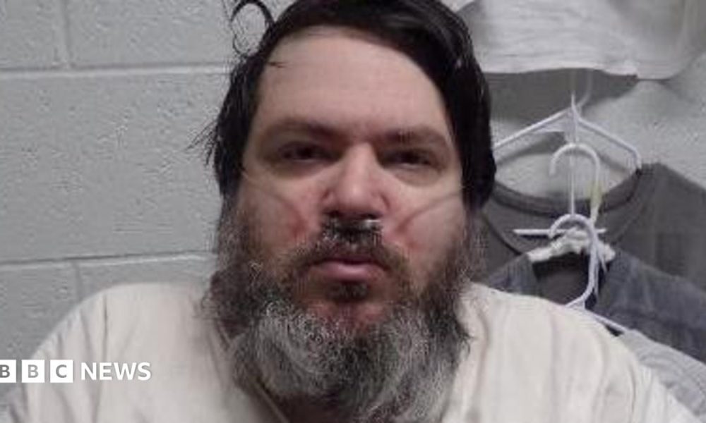A man with black hair and a long black/grey beard has a breathing tube in his nose