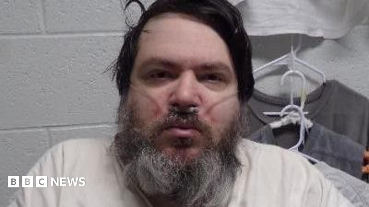A man with black hair and a long black/grey beard has a breathing tube in his nose