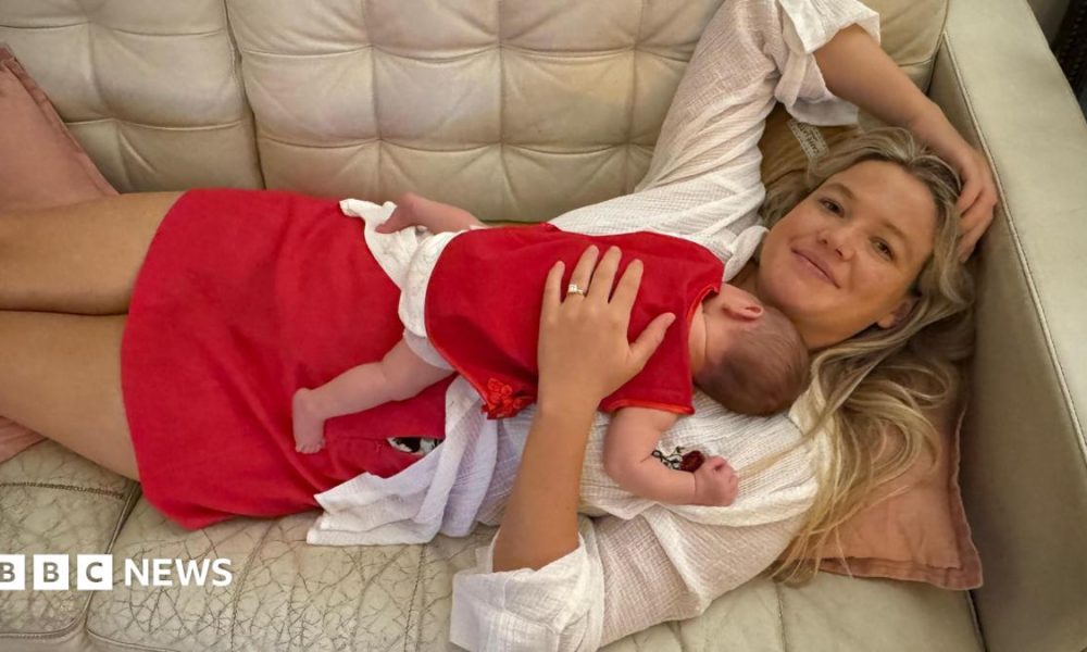 A woman with long blonde hair, wearing a white top and red skirt lying on a cream couch, with a baby sleeping on her chest.