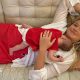 A woman with long blonde hair, wearing a white top and red skirt lying on a cream couch, with a baby sleeping on her chest.