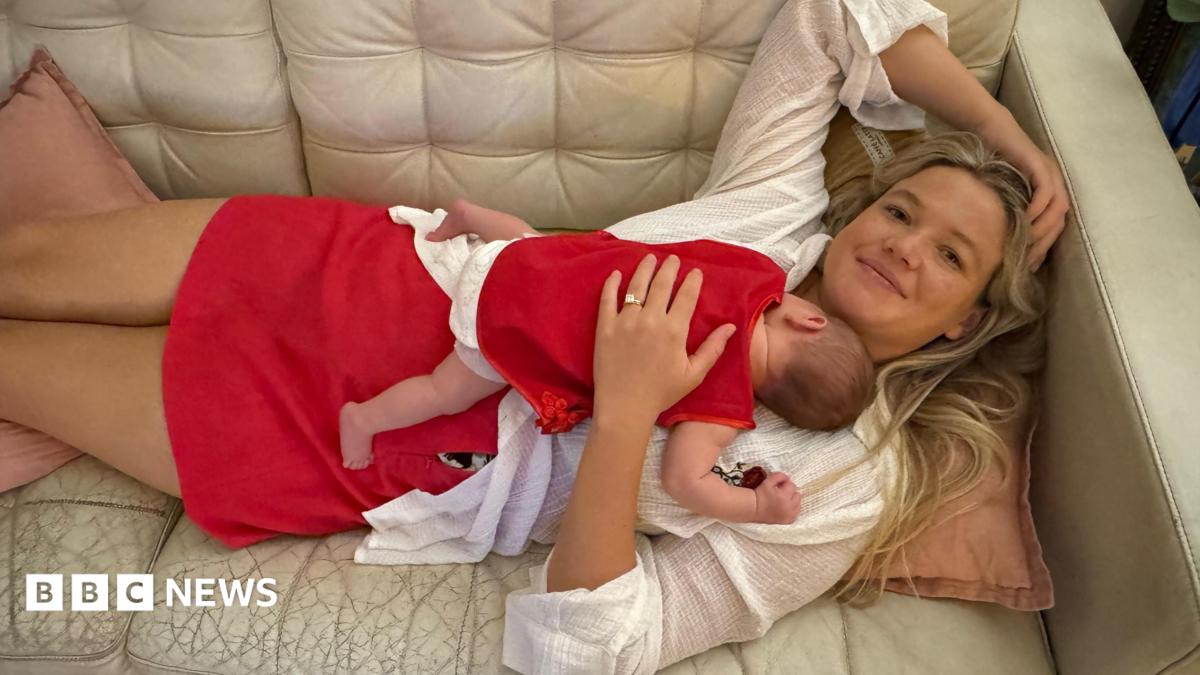 A woman with long blonde hair, wearing a white top and red skirt lying on a cream couch, with a baby sleeping on her chest.