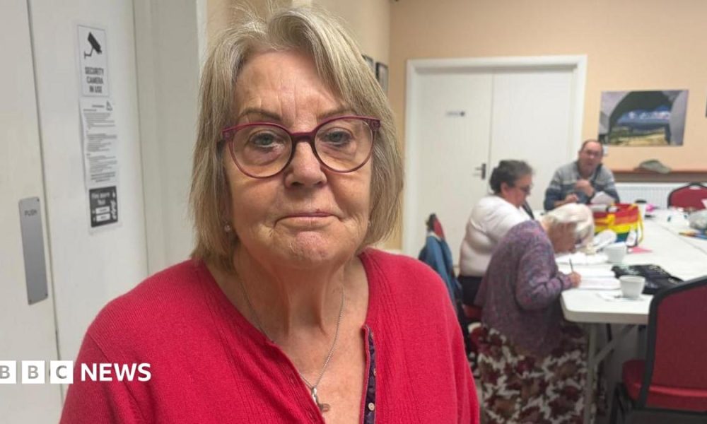 A woman with medium length hair is wearing red framed glasses and a red cardigan. She is standing in a community centre where three people are sat at a white table in the background.