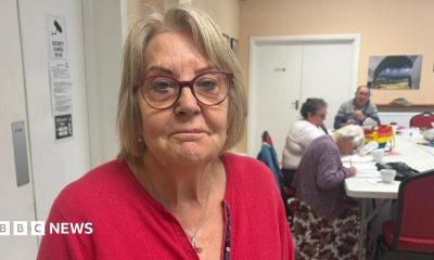 A woman with medium length hair is wearing red framed glasses and a red cardigan. She is standing in a community centre where three people are sat at a white table in the background.