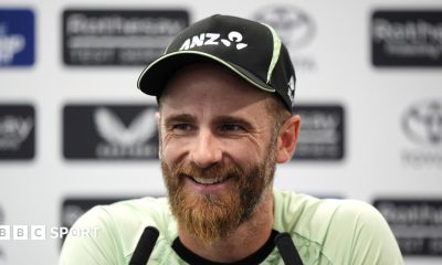 Kane Williamson smiling and wearing a black New Zealand cap sits in front of two long black microphones