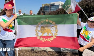 Two women wear t shirts and caps featuring the stylised lion symbol of the flag of Iran's pre-1979 Islamic revolution. They are also holding the flag between them.