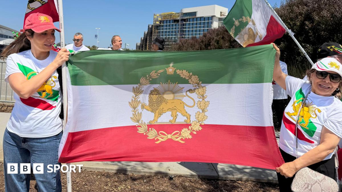 Two women wear t shirts and caps featuring the stylised lion symbol of the flag of Iran's pre-1979 Islamic revolution. They are also holding the flag between them.
