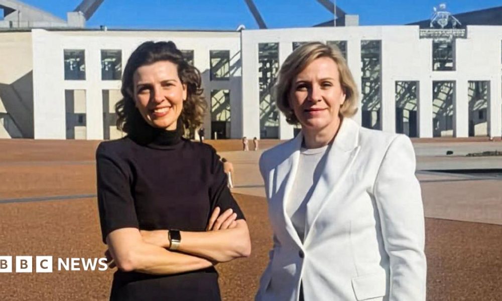 Two women standing outside federal parliament house in Canberra. One is a brunette wearing a black top and with her hands folded. The other has a short blonde bob and wearing a white blazer.