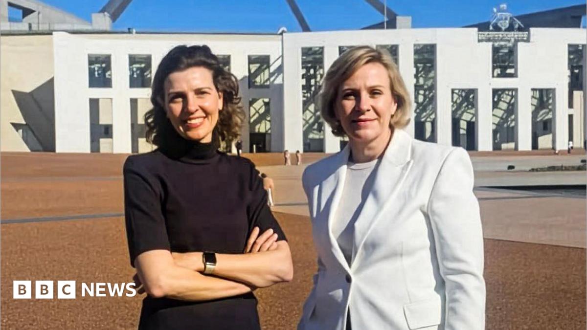 Two women standing outside federal parliament house in Canberra. One is a brunette wearing a black top and with her hands folded. The other has a short blonde bob and wearing a white blazer.