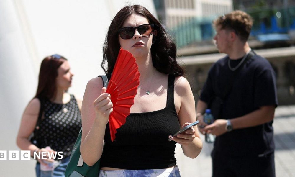 A woman on the street waves a red fan and holds her phone. She wears sunglasses and a black top.