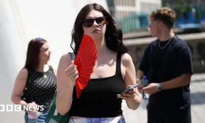 A woman on the street waves a red fan and holds her phone. She wears sunglasses and a black top.